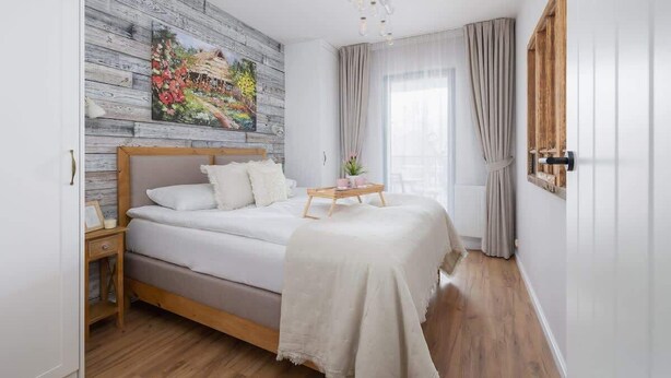 A modern bedroom with a wooden headboard, a large bed, a bedside table, a nightstand, a painting on the wall, and a window with curtains. - Stylish Apartment | Balcony and Private Parking (Kraków)