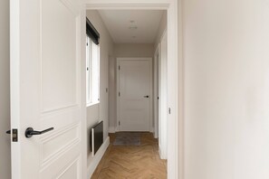 A hallway with white walls, a wooden floor, a door at the end, and a small rug on the floor.
