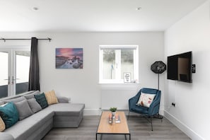 Ein modernes Wohnzimmer mit einem grauen Ecksofa, einem blauen Sessel, einem Couchtisch, einem an der Wand befestigten Fernseher und einem Fenster mit Blick auf Bäume.