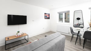 A modern living room with a flat-screen TV, a glass coffee table, a grey sofa, a dining area with a glass table and chairs, and a large mirror.