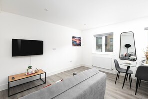 Un soggiorno moderno con una TV a schermo piatto, un tavolino in vetro, un divano grigio, un'area pranzo con un tavolo e sedie in vetro e un grande specchio.