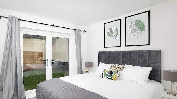 A bedroom with a bed, a nightstand, a lamp, a window with curtains, and two framed botanical prints on the wall.