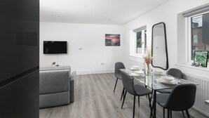 A modern dining area with a glass table, grey chairs, a flat-screen TV, and a cityscape painting.