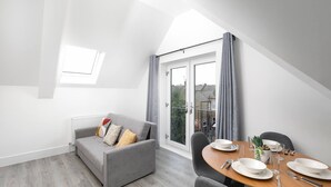 A modern living room with a grey sofa, a dining table set with plates and cutlery, and a view through a large sliding glass door. - Skyvillion -vineyard Court- Enfield 1bed Wbalcony (Enfield)