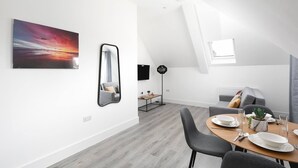 A modern dining area with a wooden table set for two, a grey sofa, and a large painting of a sunset. - Skyvillion -vineyard Court- Enfield 1bed Wbalcony (Enfield)