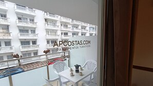 A balcony with white chairs and a table, a view of a white building with balconies, and a sign reading "APCOSTAS.COM PARAMENTOS TURISTICOS".