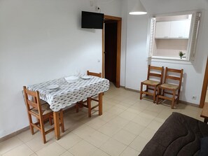 En matplats med ett bord för fyra personer, trästolar, en tv som är monterad på väggen och ett fönster med persienner.
