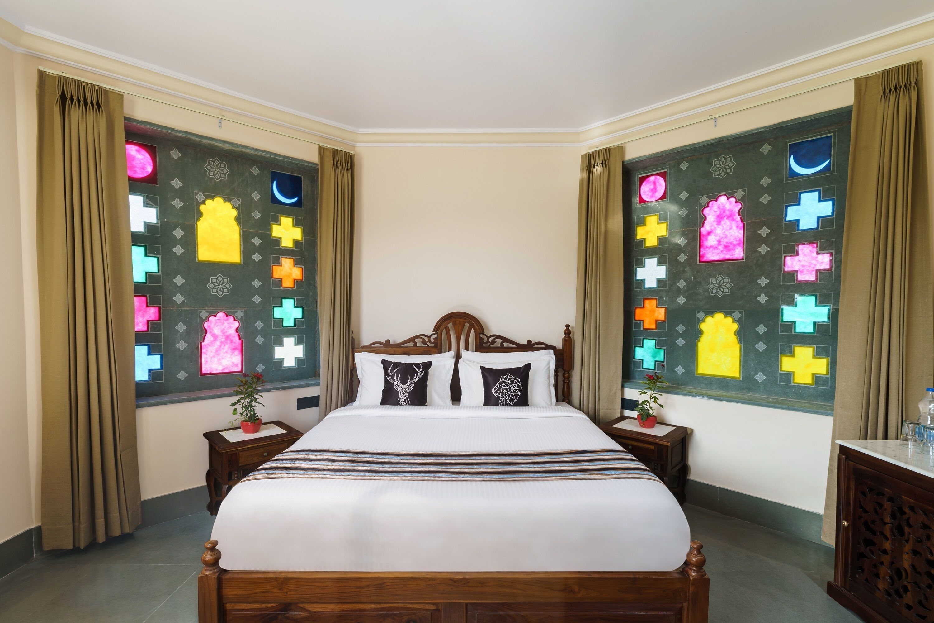 A neatly made bed with a wooden headboard, two bedside tables with potted plants, and a large window with colorful stained glass.