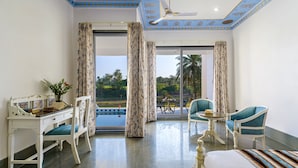 A hotel room with a desk, chair, bed, and a view of a swimming pool and palm trees. - Jhadol Bagh (Jharol)