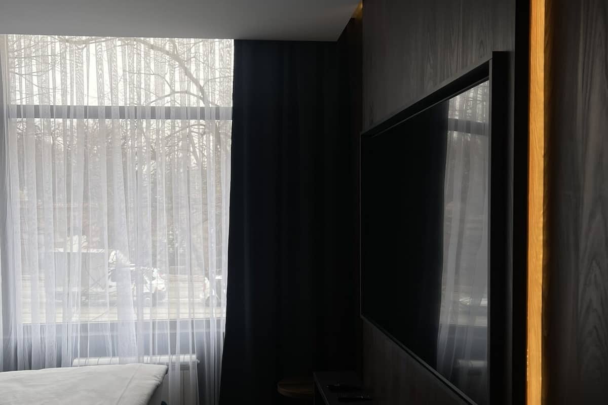 A modern hotel room with a large bed, a flat-screen TV mounted on the wall, and sheer curtains allowing natural light.
