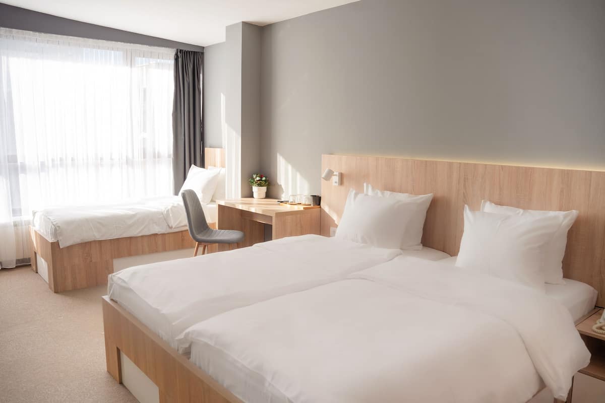 A hotel room with a large bed, a smaller bed, a desk with a chair, a nightstand, and a window with curtains.