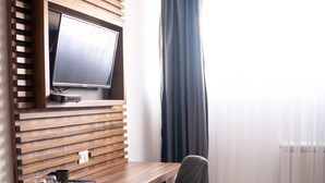 A hotel room with a wooden desk, a television mounted on a slatted wall, a chair, and a window with curtains. - Olivia Rooms (Belgrade)