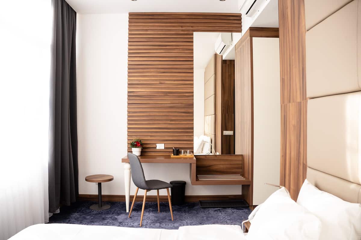 A modern hotel room with a wooden desk, a chair, a small round table, a bed, and a large mirror.