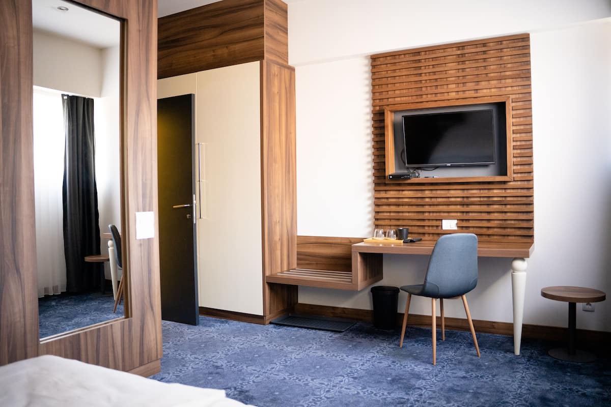 A hotel room with a wooden desk, a TV mounted on the wall, a chair, a small table, and a mirror.
