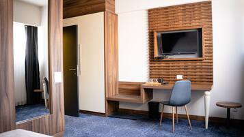 A hotel room with a wooden desk, a TV mounted on the wall, a chair, a small table, and a mirror.