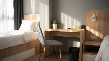 A hotel room with a bed, a desk, a chair, a small potted plant, a trash bin, and a nightstand.