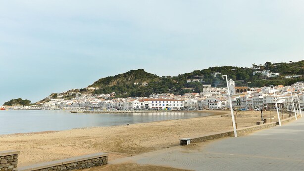 Un pueblo costero con una playa arenosa, un muro de piedra y un paseo pavimentado que lleva a una colina con edificios.