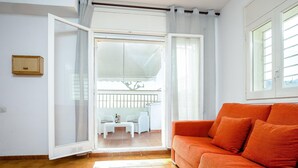 Una habitación con suelo de baldosas, un sofá blanco y vistas a un balcón con barandilla a través de una puerta abierta.