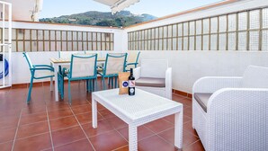 Una terraza con mobiliario blanco de mimbre, una mesa y sillas, con vista a un paisaje montañoso.