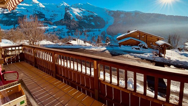 Une cabane en bois, dont le toit est recouvert de neige, entourée d’une terrasse en bois et avec une chaîne de montagnes en arrière-plan.