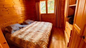 Une chambre confortable dans un chalet, avec un intérieur en bois, un lit recouvert d’une couverture moelleuse, deux oreillers, une fenêtre avec des rideaux et une commode avec un miroir.