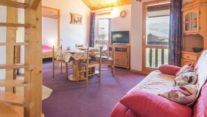 Ein gemütliches Zimmer mit Holzbalkendecke, einem Essbereich mit Tisch und Stühlen, einem Fernseher und einem großen Fenster mit Blick ins Freie.