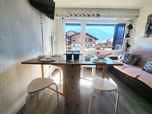 A dining area with a wooden table, two white chairs, and a sofa with patterned cushions.