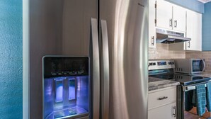 Un refrigerador de acero inoxidable con dispensador de agua, un microondas y una estufa en una cocina con armarios blancos y pared azul.