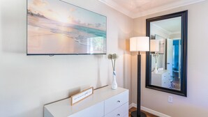 Una habitación con un televisor de pantalla plana montado en la pared, un aparador blanco con cajones, una lámpara de pie y un espejo grande.