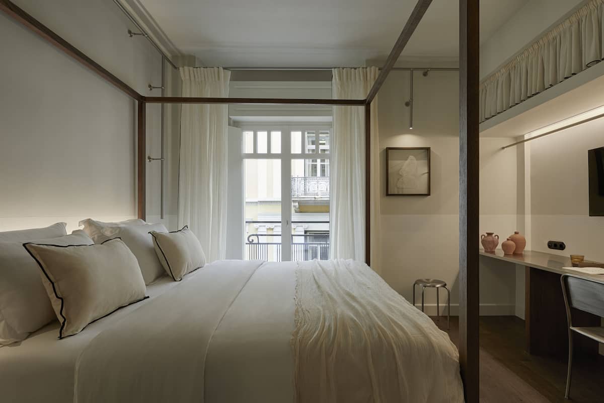 Habitación de hotel con cama, escritorio, silla, taburete pequeño y vista al balcón exterior.