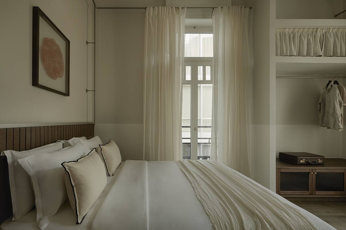 Un dormitorio ordenado con cama, almohadas, un cuadro enmarco, una ventana con cortinas y un armario con ropa colgada.