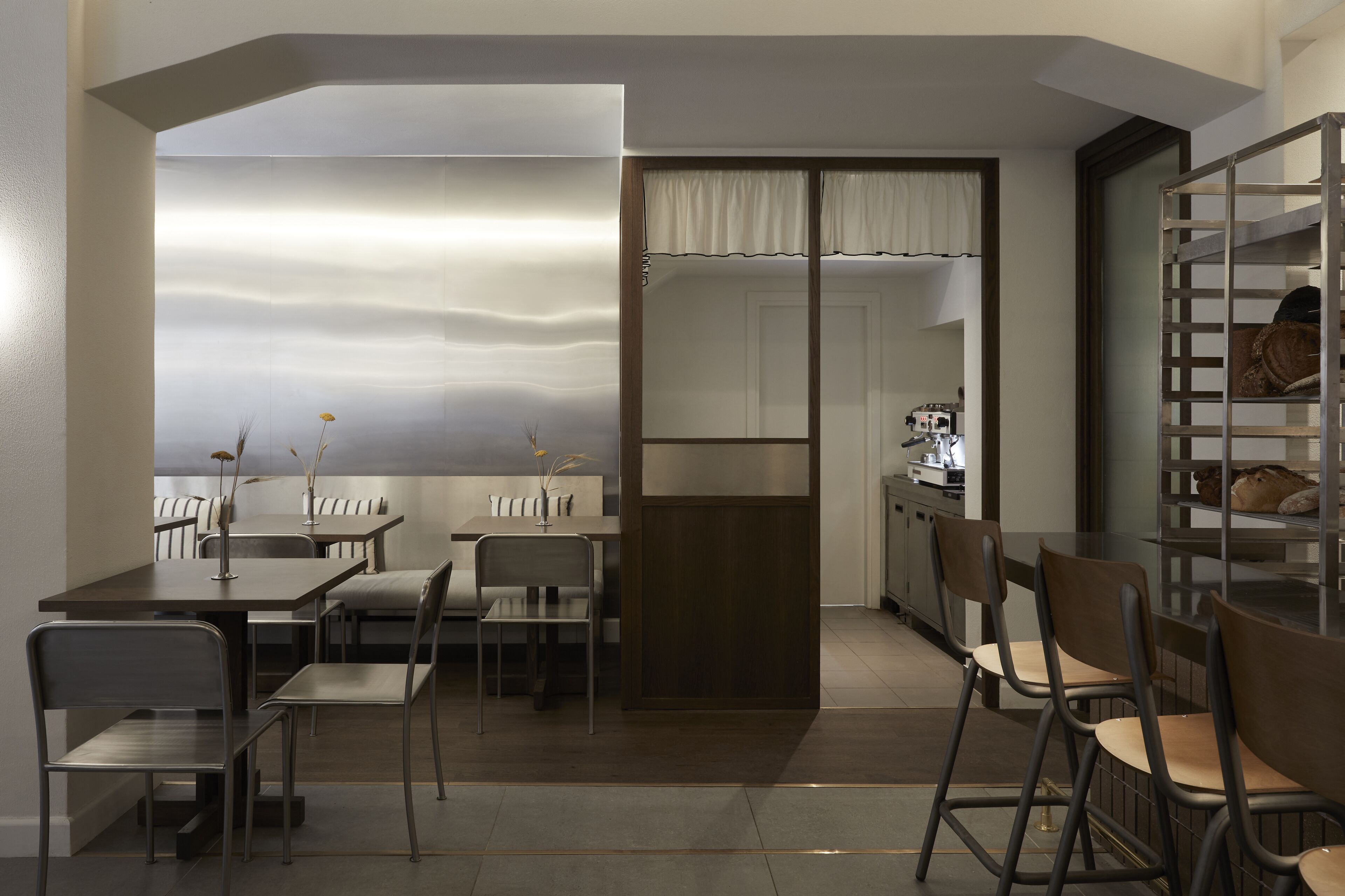 A modern dining area with a long table and chairs, a kitchen in the background, and a view through a glass partition.