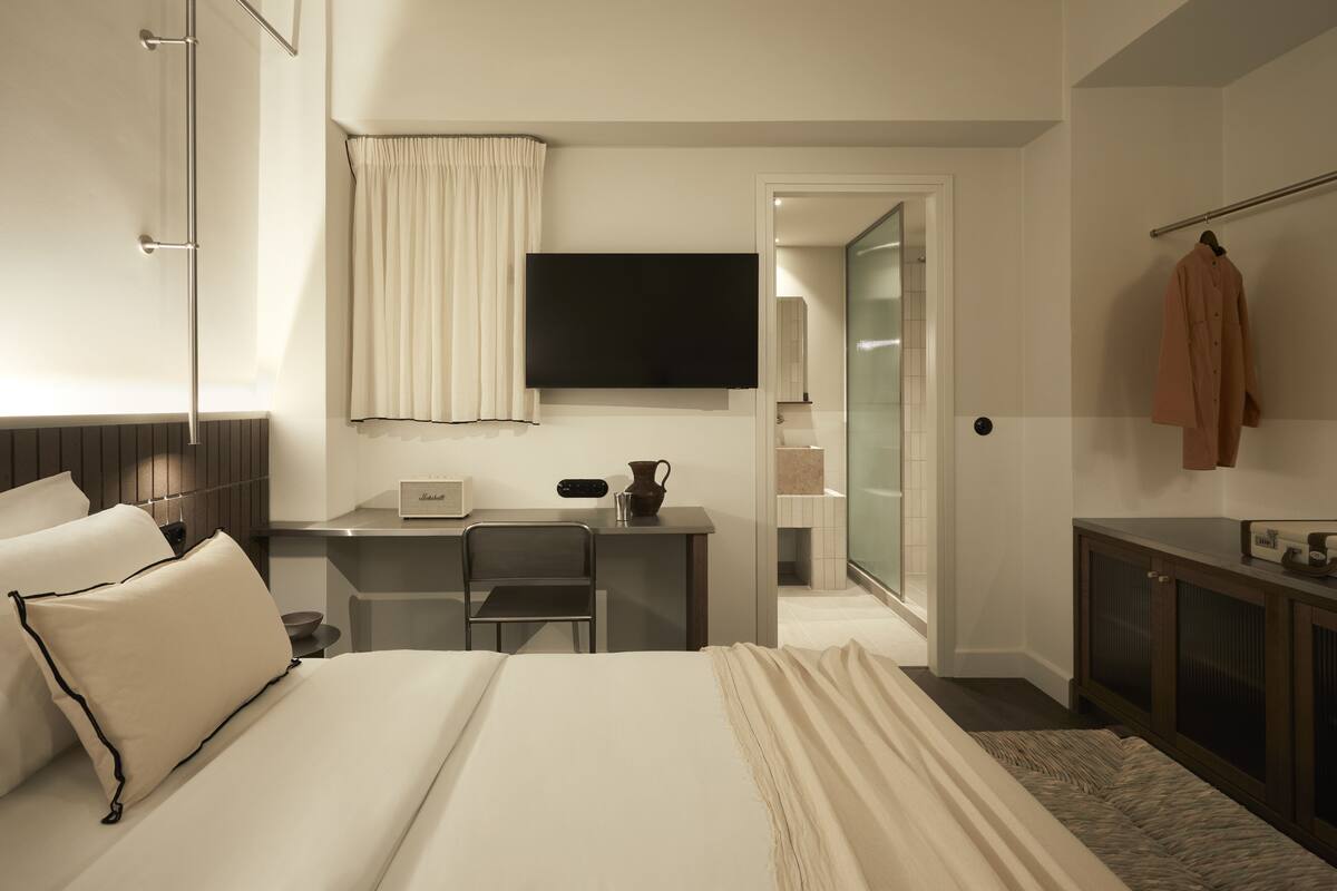 Habitación de hotel con cama, escritorio, silla, televisor, armario y baño visible a través de una puerta abierta.