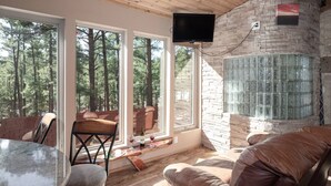 A cozy room with a stone fireplace, leather seating, and large windows overlooking a forest. - Sandoval in the Historic Upper Canyon (Ruidoso)