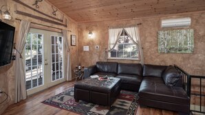 A cozy living room with a leather sofa, a coffee table, and a wooden floor. There is a TV mounted on the wall, a window with curtains, and a painting on the wall. - Sandoval in the Historic Upper Canyon (Ruidoso)