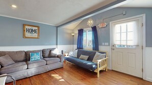 A cozy living room with a grey sofa, a blue sofa, and a wooden bed. There is a framed sign on the wall that says "HOME is where the waves crash." - Gull's Nest by Bayley Vacation Rentals (Old Orchard Beach)