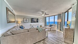 A living room with a large sofa, a smaller sofa, a coffee table, and a cabinet. There is a ceiling fan, a window with a view of the ocean, and artwork on the wall. - Tower 2-1002 Edgewater Beach Resort-Sleeps 8! (Panama City Beach)