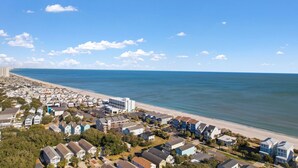 Vue aérienne d’une zone résidentielle côtière, avec des maisons situées près de la plage et un ciel bleu dégagé.