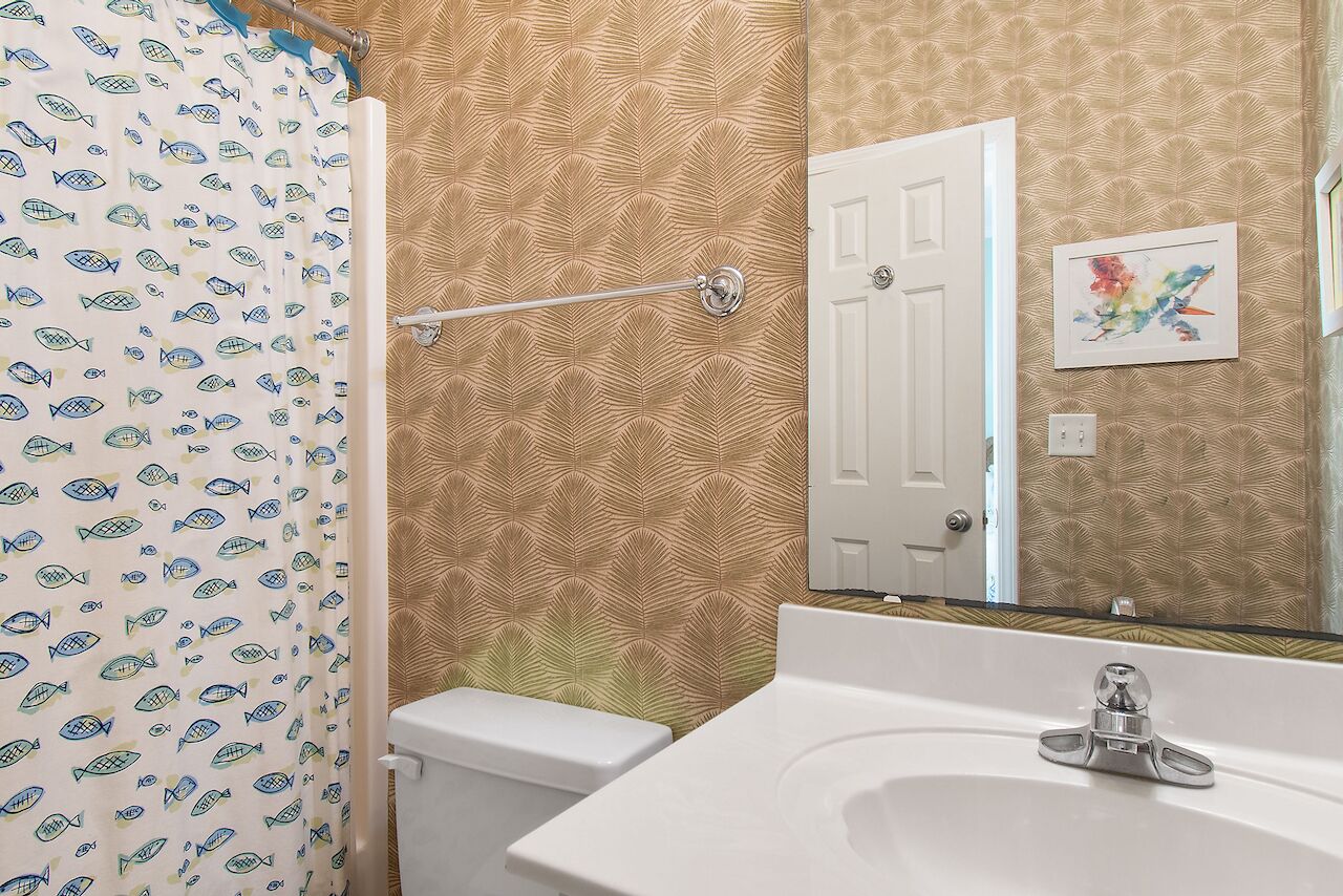 Une salle de bain avec un rideau de douche orné de motifs de poissons, des toilettes blanches, un lavabo avec un robinet moderne et une porte agrémentée d’un miroir décoratif.
