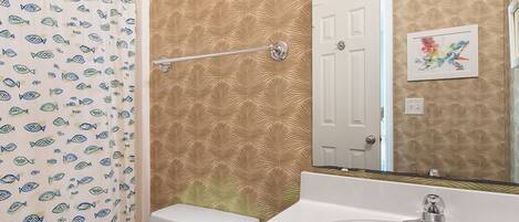 Une salle de bain avec un rideau de douche orné de motifs de poissons, des toilettes blanches, un lavabo avec un robinet moderne et une porte agrémentée d’un miroir décoratif.