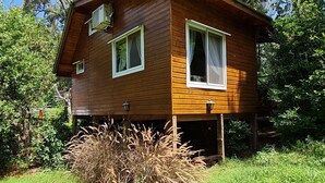 Uma cabana de madeira com duas janelas, rodeada por vegetação e árvores.