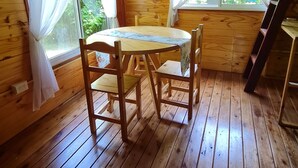 Interior de uma cabana de madeira com uma mesa redonda e cadeiras, duas janelas com cortinas e um ventilador de teto.
