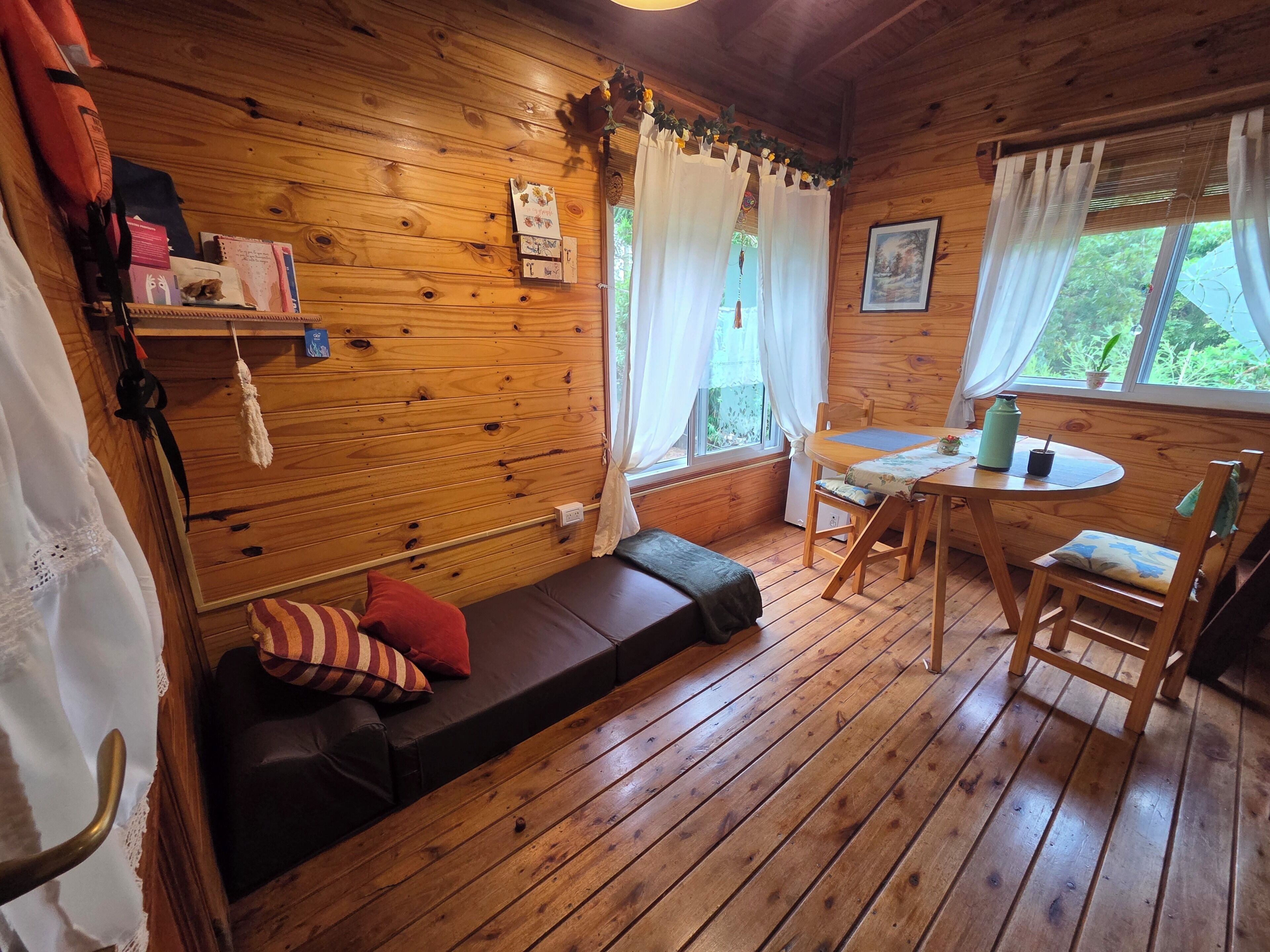 A wooden cabin interior with a sofa, a dining table, and a window with curtains.
