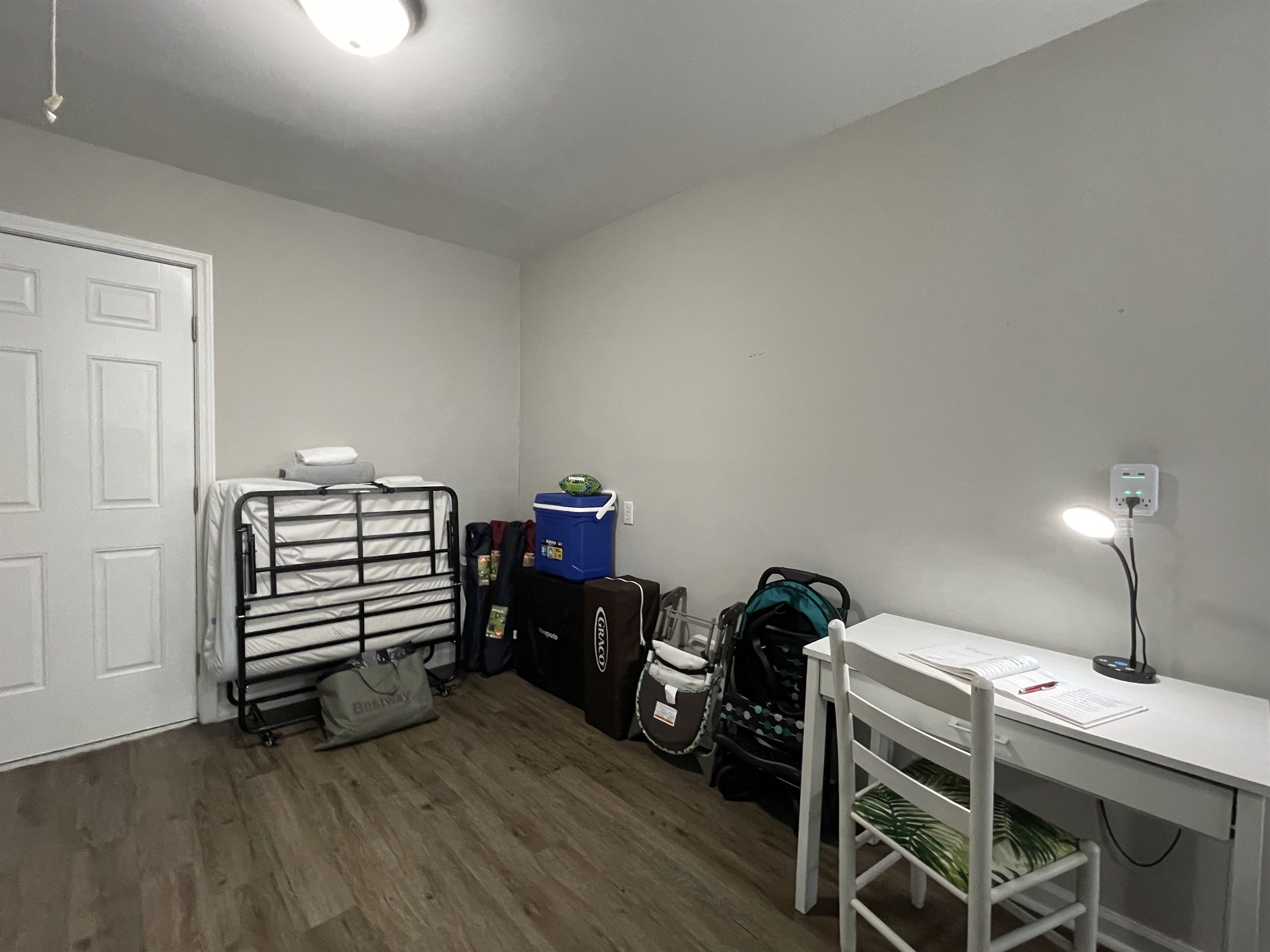 A room with a white desk, a chair, a bed with a metal frame, a suitcase, a backpack, and a bag on the floor.