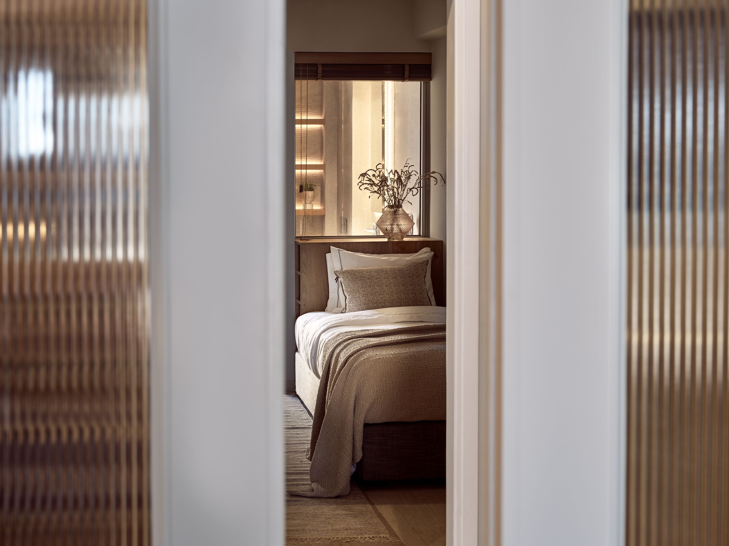 A bedroom with a bed, a bedside table with a vase, and a window with blinds.
