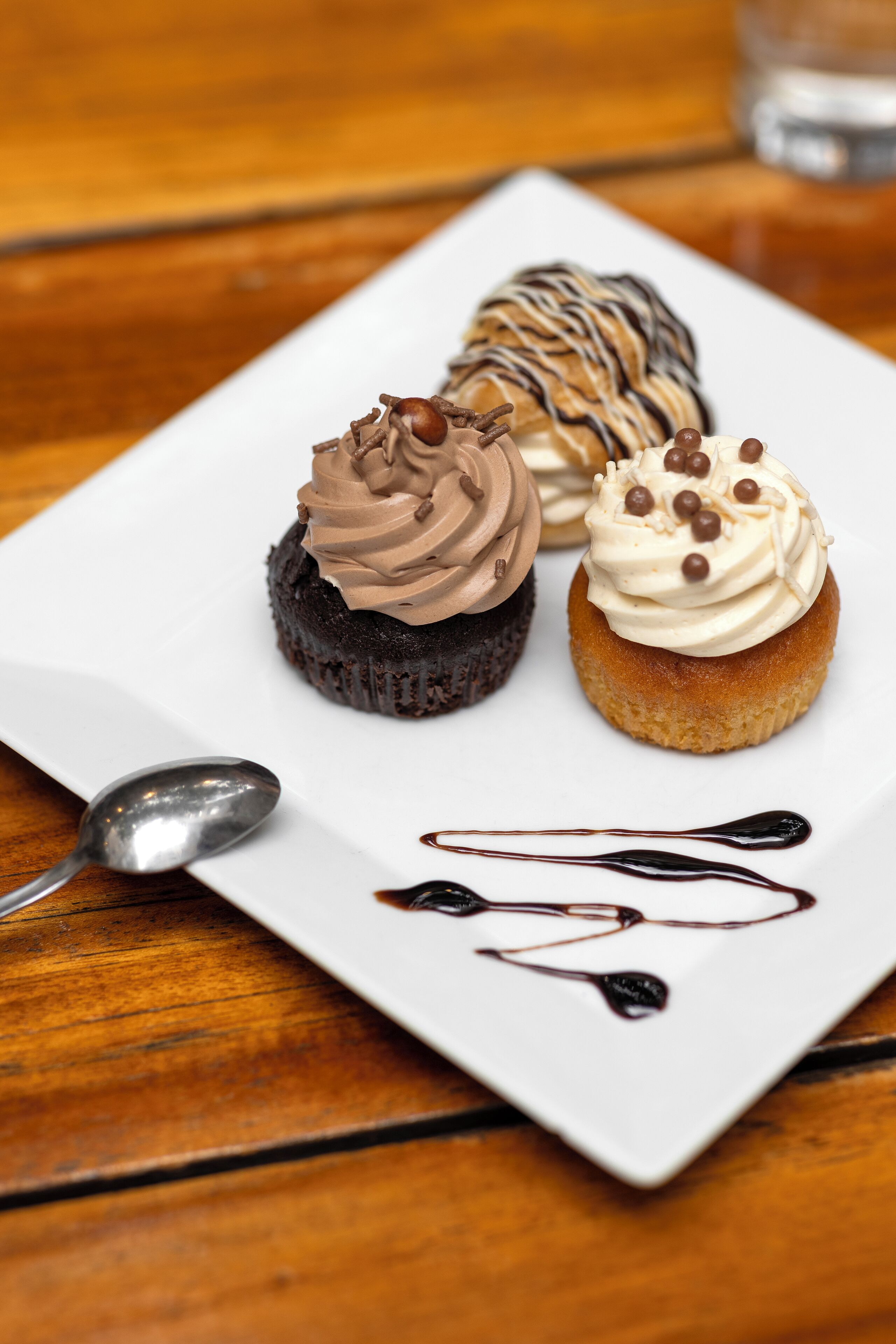 A dessert plate with three cupcakes on a white square plate, a spoon, and chocolate syrup on a wooden table.