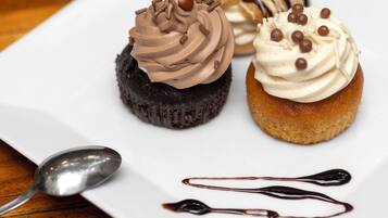 Un plato con tres cupcakes sobre una fuente cuadrada blanca, una cuchara y almíbar de chocolate sobre una mesa de madera.