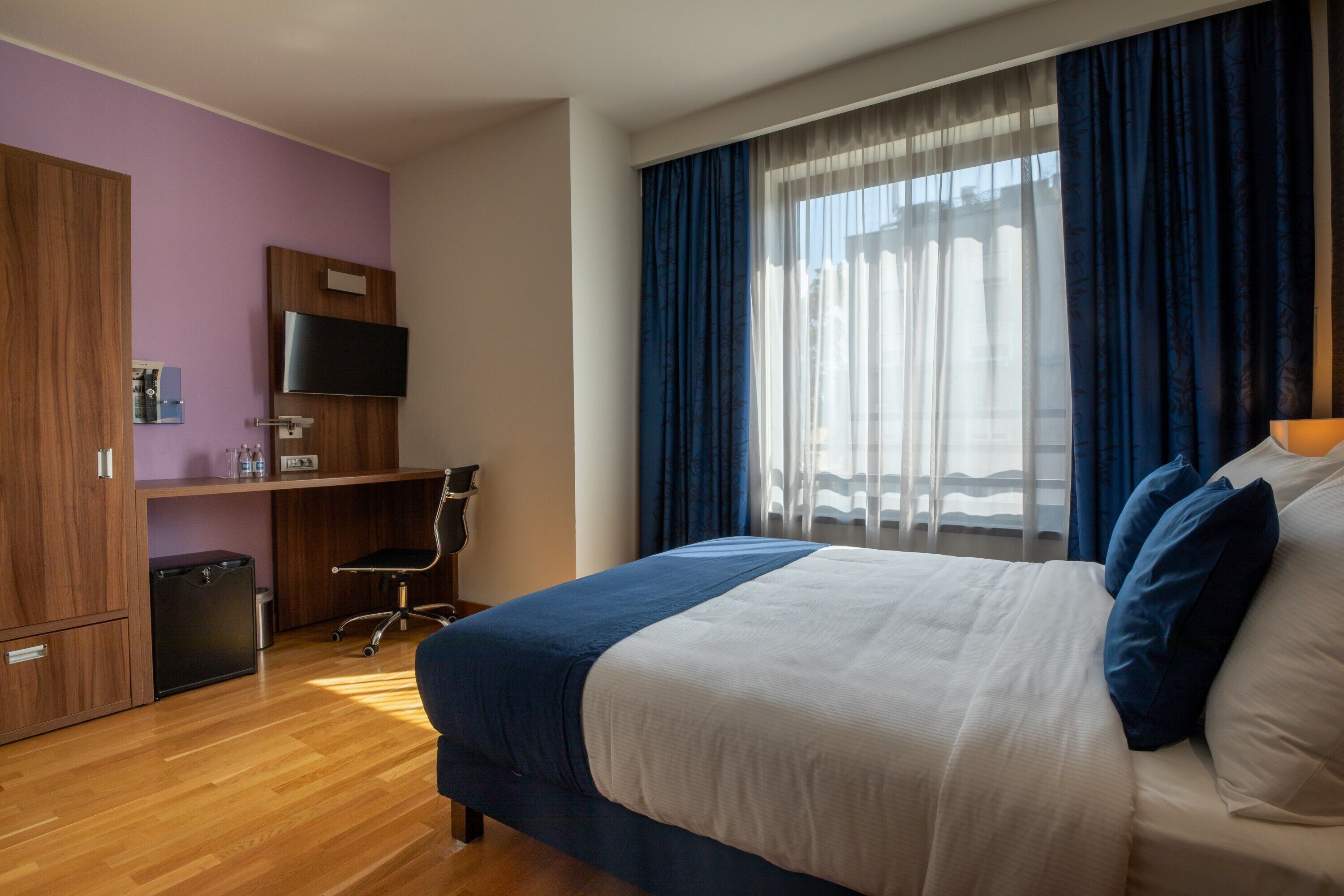 Une chambre d’hôtel avec un grand lit, un bureau avec un ordinateur, une télévision et une fenêtre avec des rideaux.