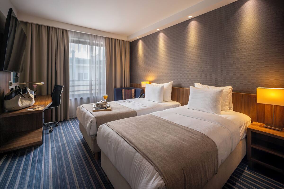 A hotel room with two beds, a desk with a chair, a TV, and a window with curtains.