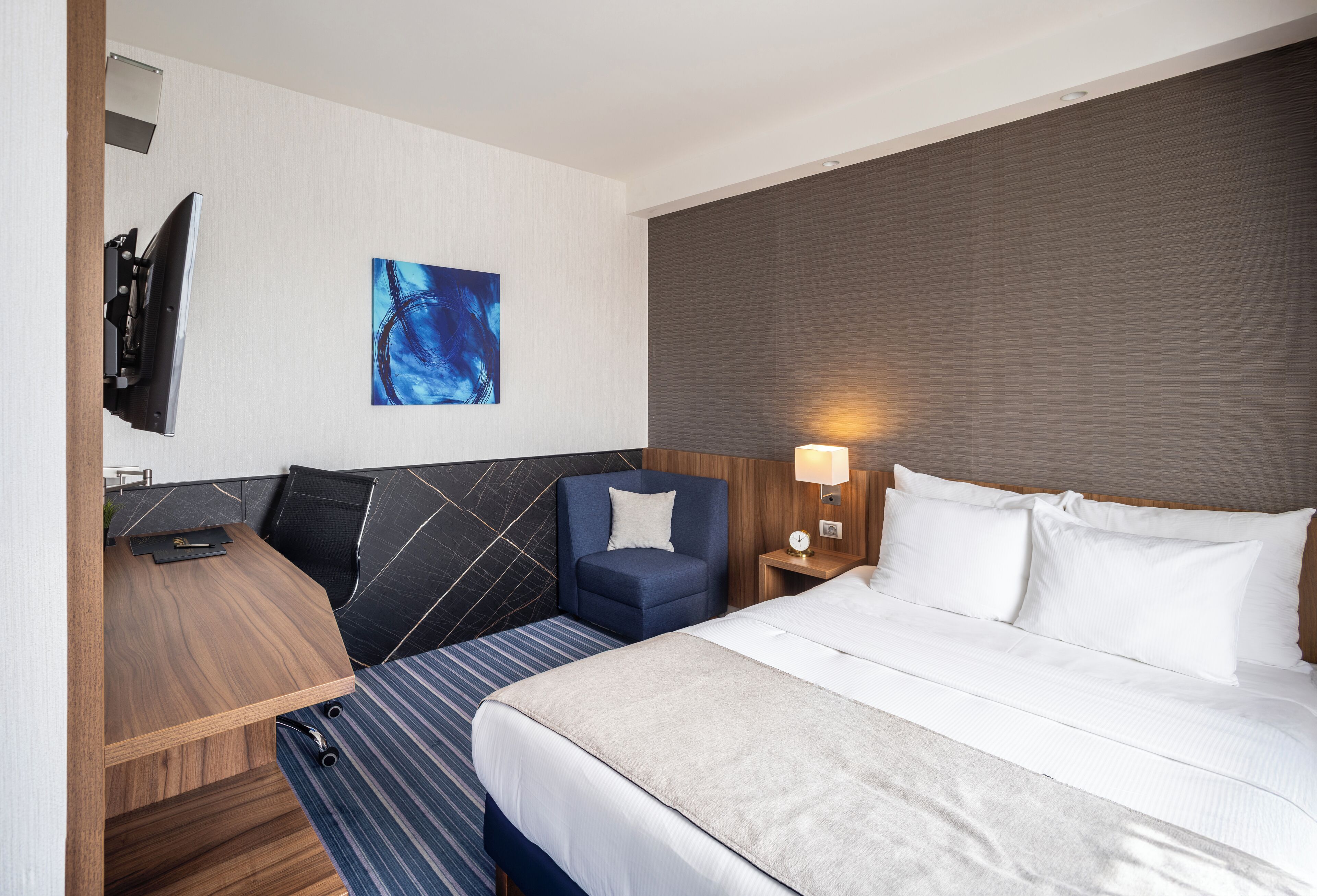 A hotel room with a bed, a desk, a chair, a TV, and a painting on the wall.
